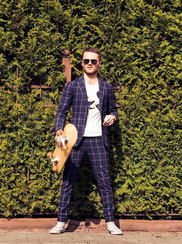 Emotional handsome bearded man in sunglasses and classical suit poses with skateboard on green bush background.Fashion look.Sportrs guy, stock photo