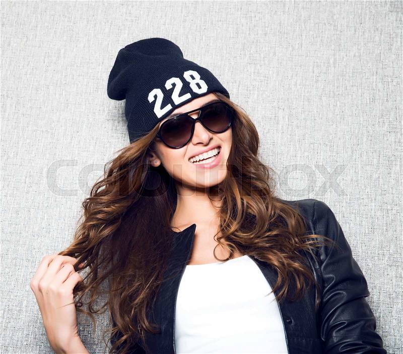 Portrait of beautiful long haired girl in stylish sunglasses,black cap,white shirt,black jacket and jeans poses on grey background.Fashion look.Stylish image.Casual.Lifestyle.Studio shot.Dj woman, stock photo