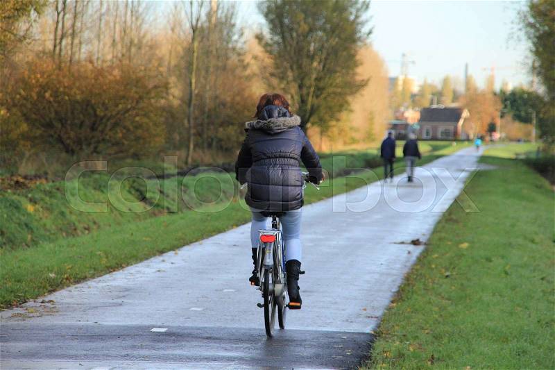 The woman is biking and in the distance a walking couple at the cycle path at the countryside in fall, stock photo
