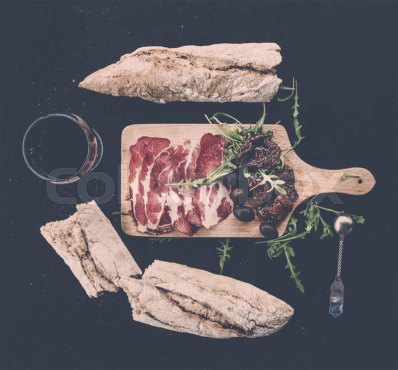 Wine appetizer set: vintage dinnerware, french baguette broken into pieces, dried tomatoes, olives, smoked meat and arugula on rustic wooden board over dark background. Top view, pastel tone, stock photo