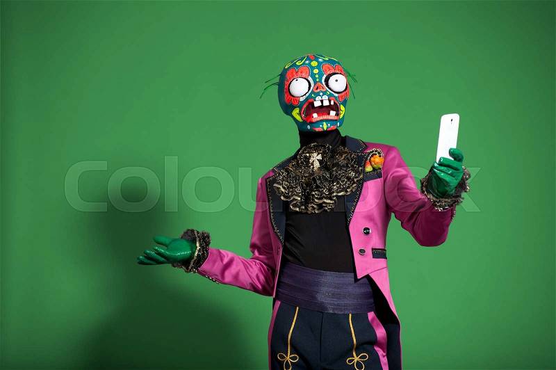 Actor in pink suit and zombie mask poses on green background taking selfie.Halloween party time.Spooky man.Studio shot, stock photo