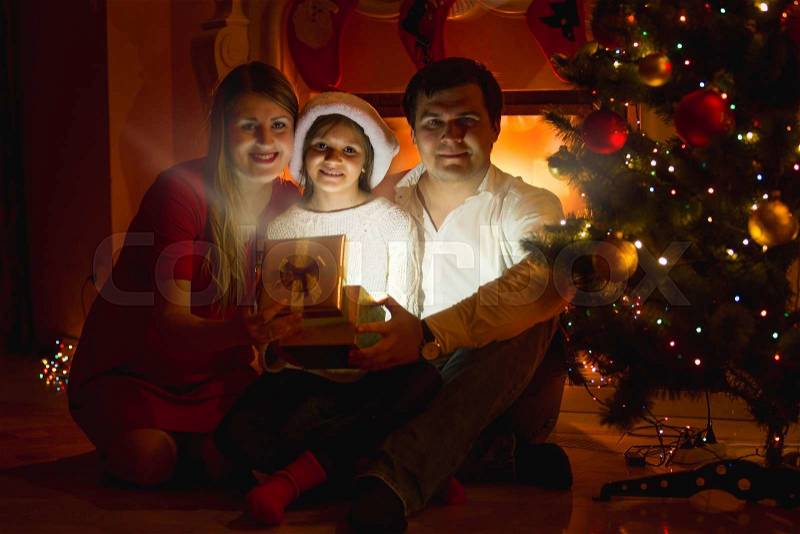 Happy smiling family sitting at fireplace and opening Christmas gift box, stock photo