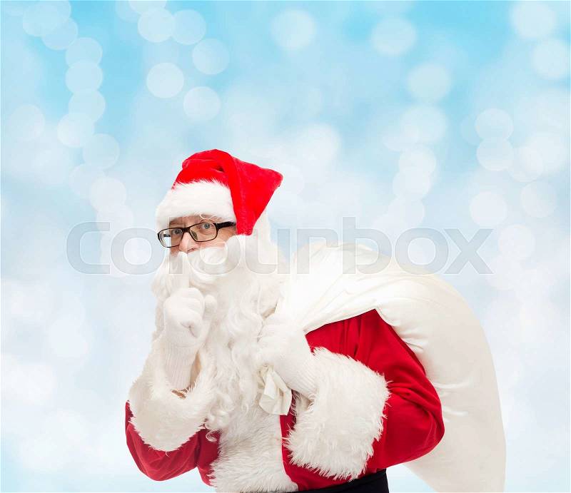 Christmas, holidays and people concept - man in costume of santa claus with bag making hush gesture over blue lights background, stock photo