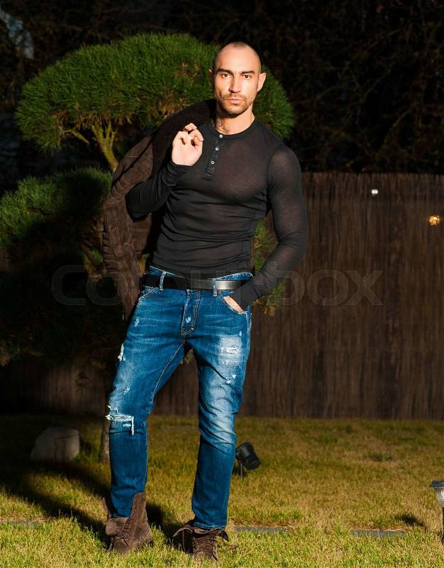 Attractive young muscular man in brown leather jacket, black shirt, blue jeans poses outside at night.Fashion look.Bearded guy.Modern style, stock photo
