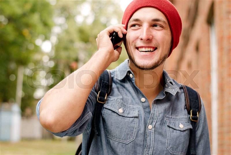 The young man with red cap calls on his cell phone on background with trees and building, stock photo
