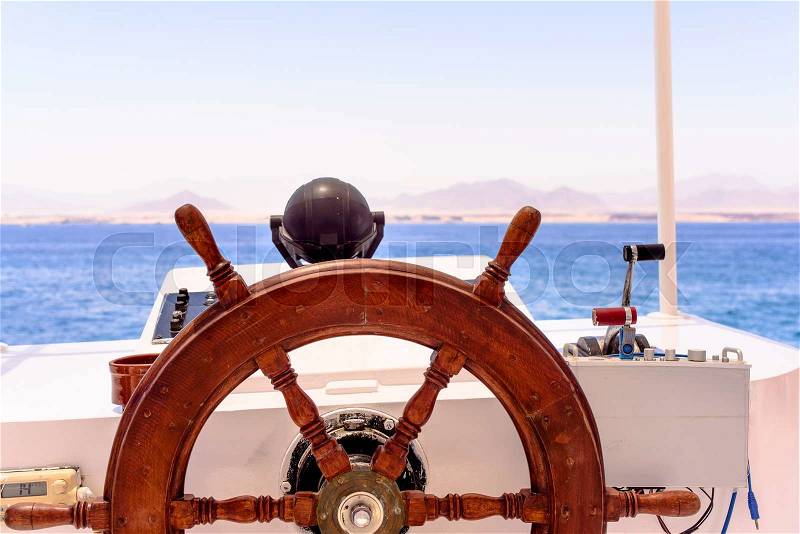 Ships wooden spooked wheel and navigation console at the helm of a yacht or motorboat cruising mid ocean with a distant coastline visible on the horizon, nobody in view, stock photo