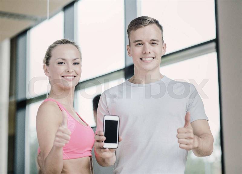 Fitness, sport, advertising, technology and diet concept - smiling young woman and personal trainer with smartphone in gym showing thumbs up, stock photo
