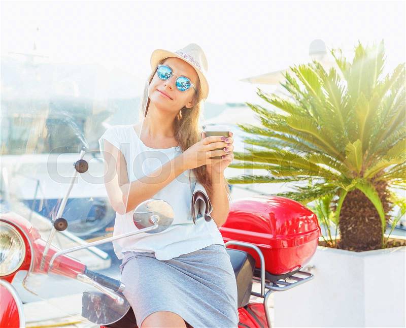 Happy young trendy woman drinking takeaway coffee near her red moped on the waterfront in the morning, stock photo