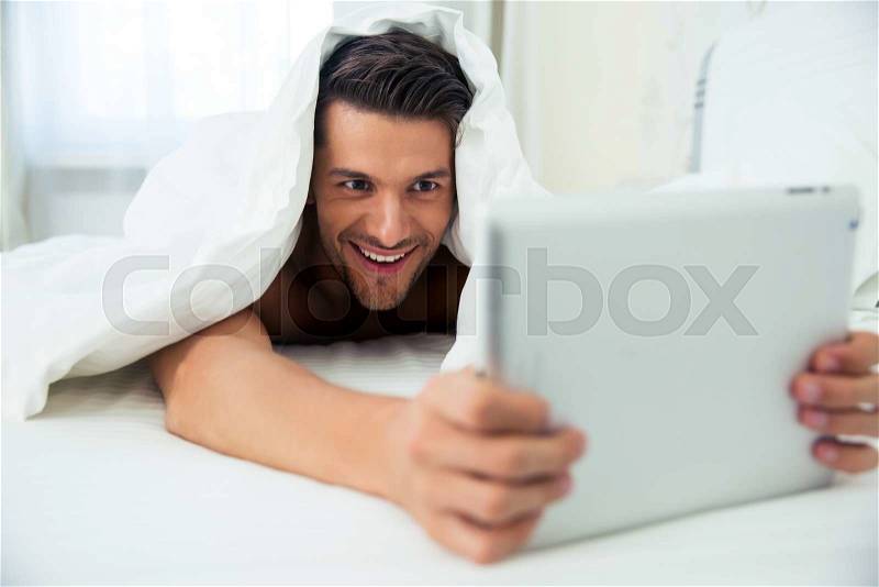 Portrait of a smiling man lying under blanket and using tablet computer at home, stock photo