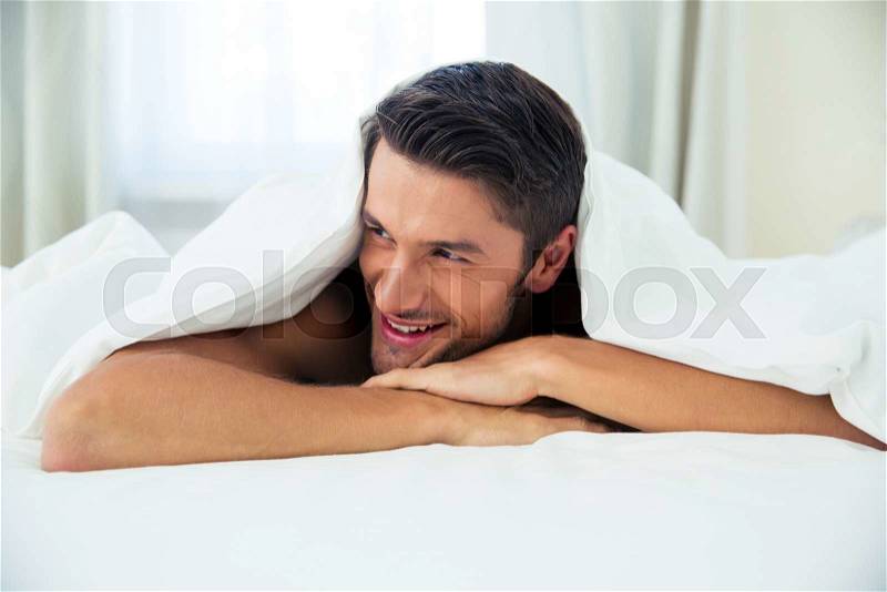 Portrait of a smiling man lying under blanket on the bed at home, stock photo
