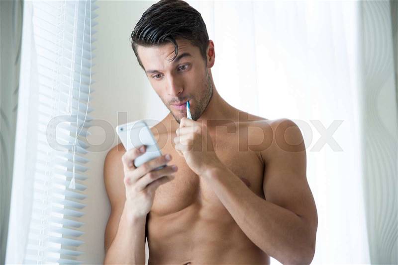Portrait of a man with fitness body brushing teeth and using smartphone at home, stock photo