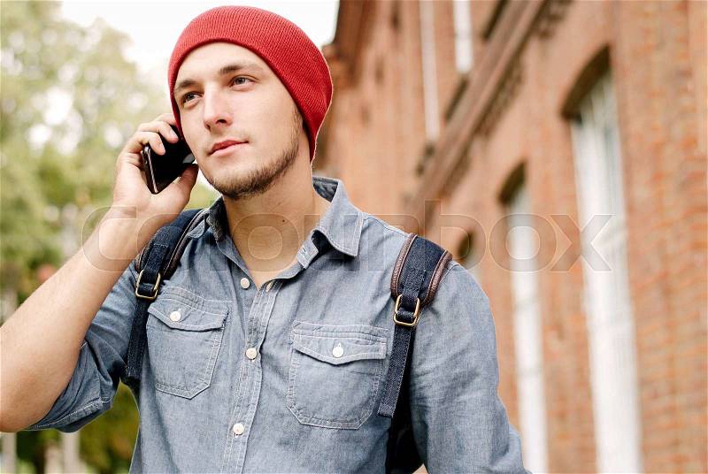 The young man with red cap calls on his cell phone on background with trees and building, stock photo