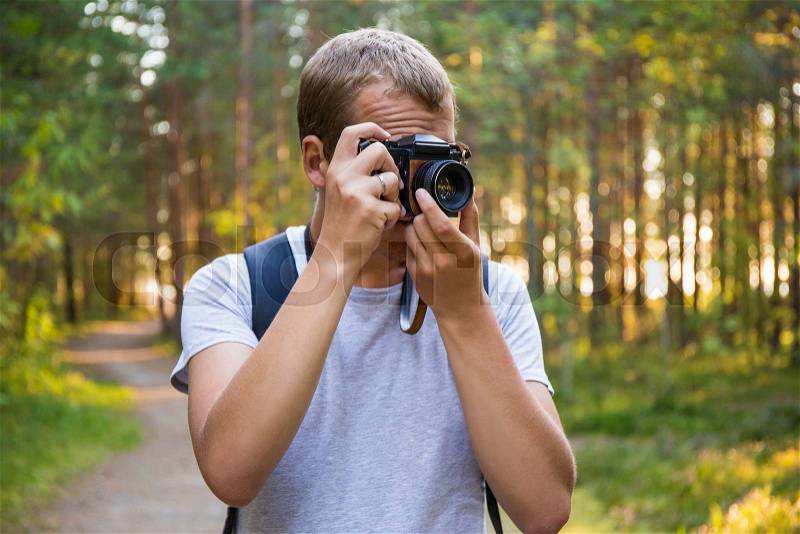 Young man with backpack taking a photo with retro camera in forest, stock photo
