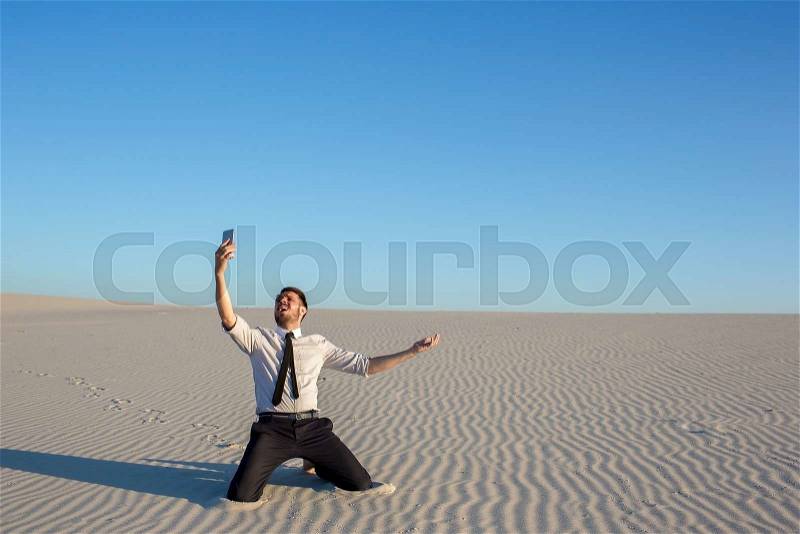 Poor signal. Frustrated young businessman searching for mobile phone signal while sitting on sand in desert, stock photo