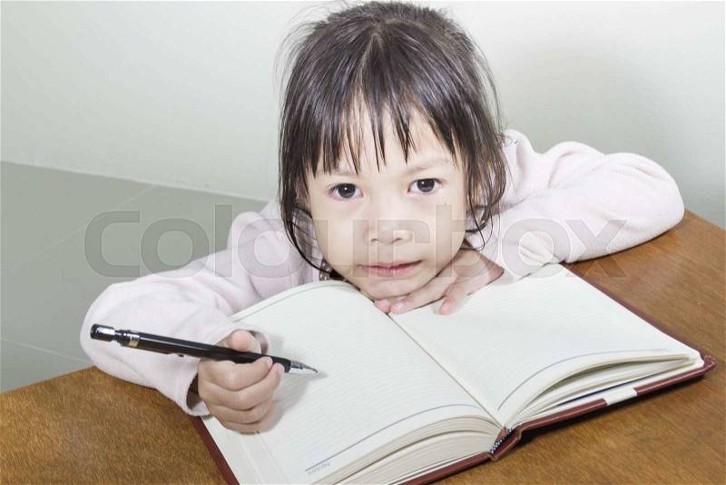 Asian girl kid hard at work in the room sitting with his head on her hand writing notes on book of white paper, stock photo