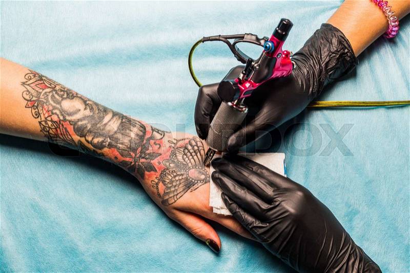 Tattooist draws black and red pattern on the clients hand and holding a tattoo machine and sterile black gloves. Tattoo artist works on a blue professional mat. , stock photo