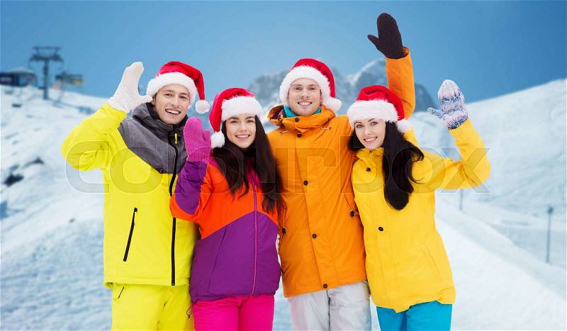 Winter holidays, christmas, friendship and people concept - happy friends in santa hats and ski suits over downhill skiing and mountains background, stock photo