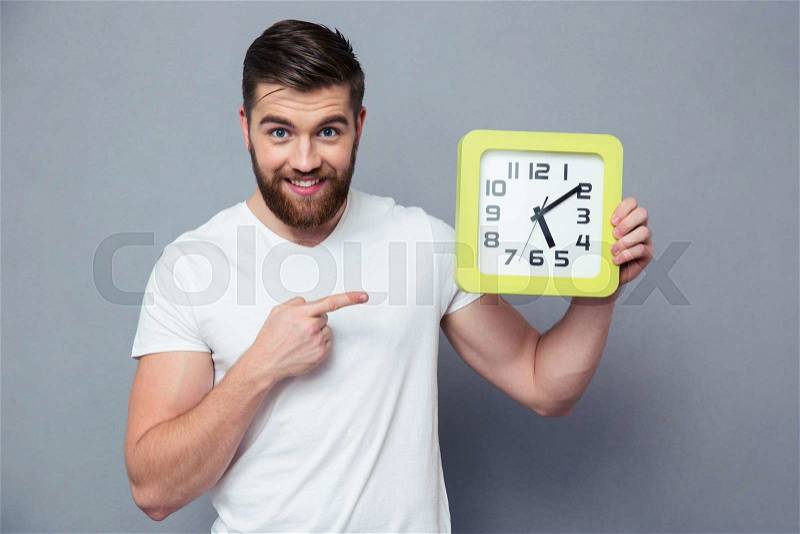 Portrait of a happy casual man pointing finger on wall clock over gray background, stock photo