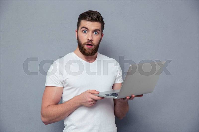 Portrait of amazed man holding laptop computer and looking at camera over gray background, stock photo