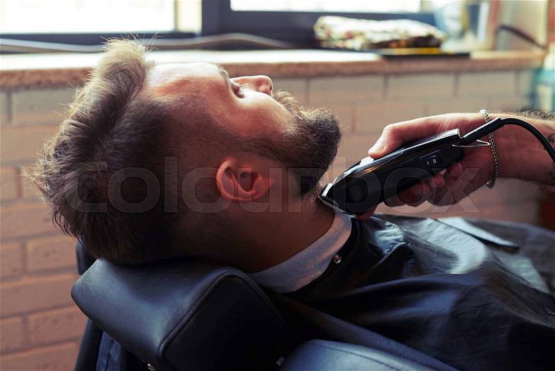 Barber shaving beard with electric razor in barber shop, stock photo