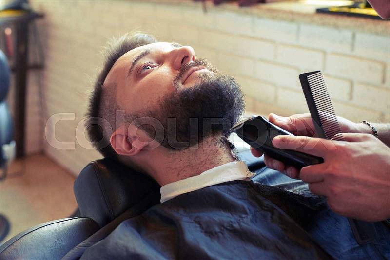 Barber shaving beard with electric razor and holding comb in barbershop, stock photo