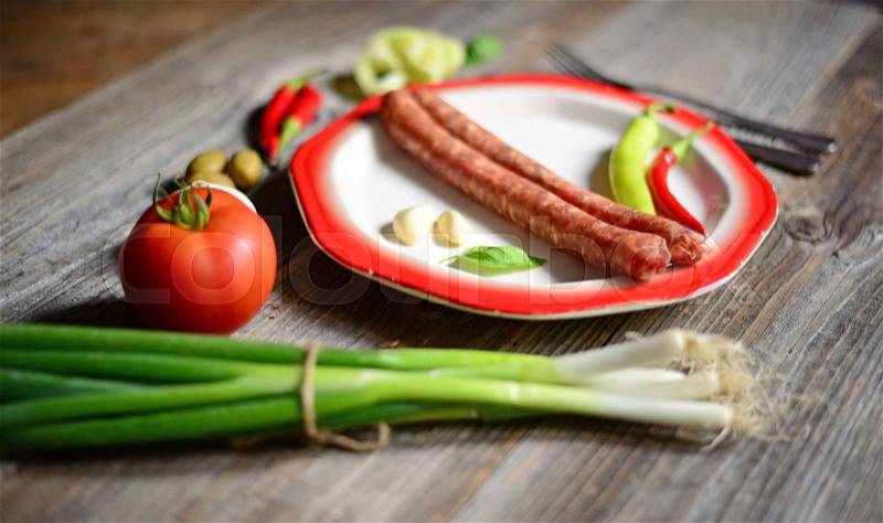 Portion of sausages on plate with tomatoe, fresh onion, chilly peppers and olives on wooden background, stock photo