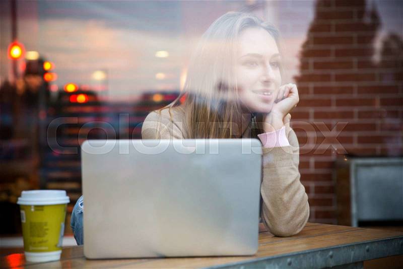 Portrait of a happy girl sitting with laptop in cafe and looking away in window, stock photo