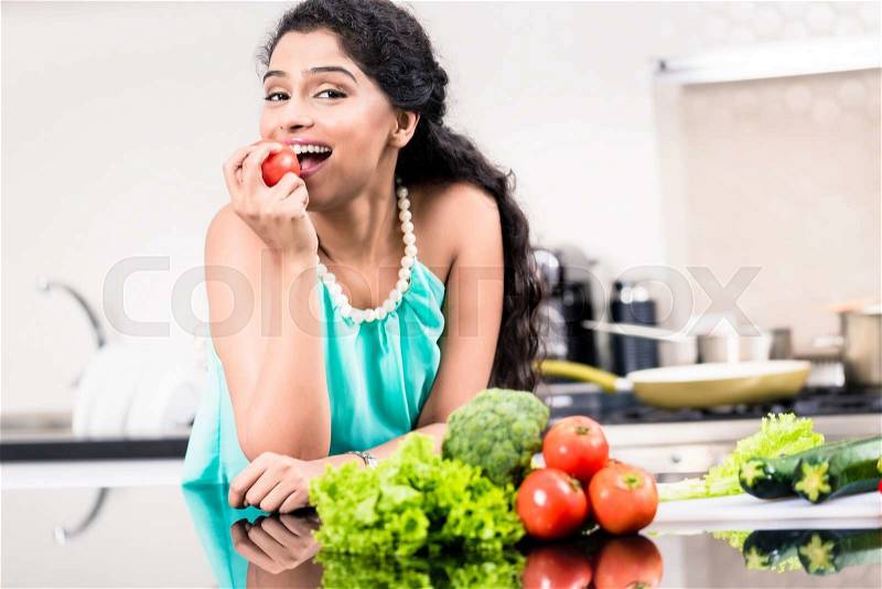 Indian woman eating healthy apple in her kitchen, salad and vegetables on the counter, stock photo