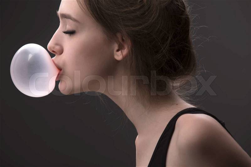 Portrait of sexual brown-haired, which inflates languidly pink bubble of chewing gum. Photographed in studio on a dark background, stock photo