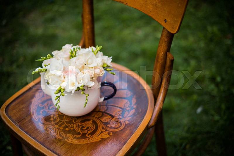 Summer wedding party festive decor, lovely fresh bouquet of white summer roses and freesias in vintage enamel tea pot on an old brown wooden chair. Retro style outdoor decoration, grass background, stock photo