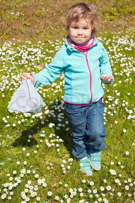 Cute Caucasian blond girl took off her hat on green lawn with daisies flowers, stock photo