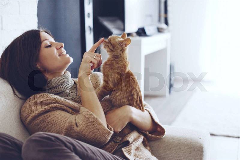 Young woman wearing warm sweater is resting with a cat on the armchair at home one autumn day, stock photo
