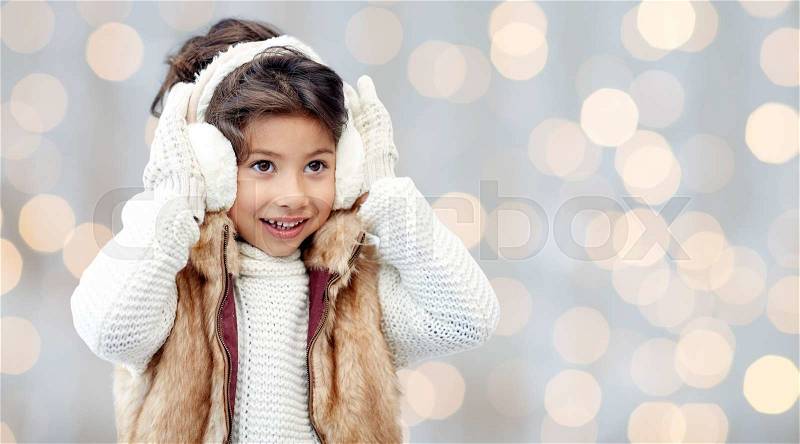 Winter, people, christmas, fashion and childhood concept - happy little girl wearing earmuffs and gloves over holidays lights background, stock photo