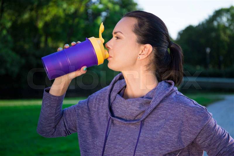 Close up of fitness woman drinking water from bottle in autumn park, stock photo