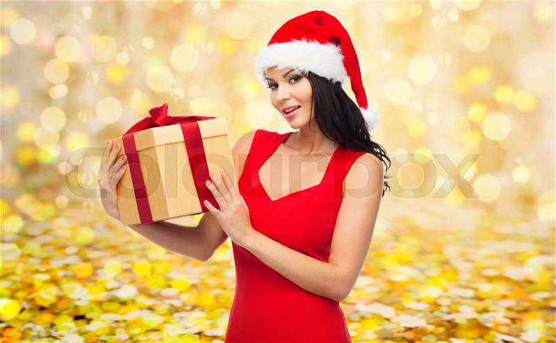 People, holidays, christmas and celebration concept - beautiful sexy woman in red dress and santa hat with gift box over yellow lights or golden confetti background, stock photo