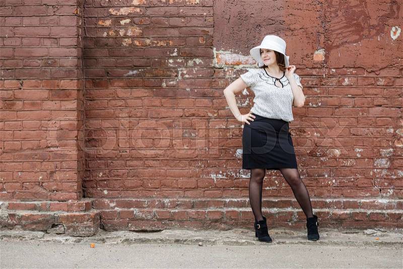 Woman in a white hat, blouse and black skirt, posing against the backdrop of an old vintage brown brick wall. Female portrait in a retro style, stock photo