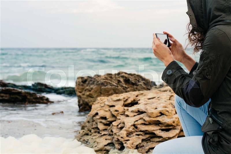 Woman in jeans and a green jacket with a hood pictures of the sea on the phone in the autumn, stock photo