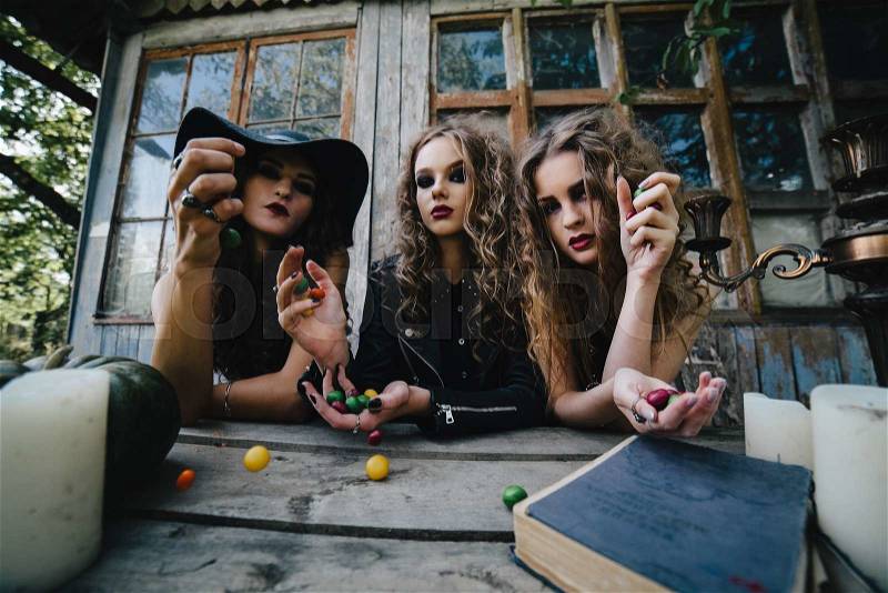 Three vintage witches perform magic ritual, throwing sweet at a table on the eve of Halloween, stock photo