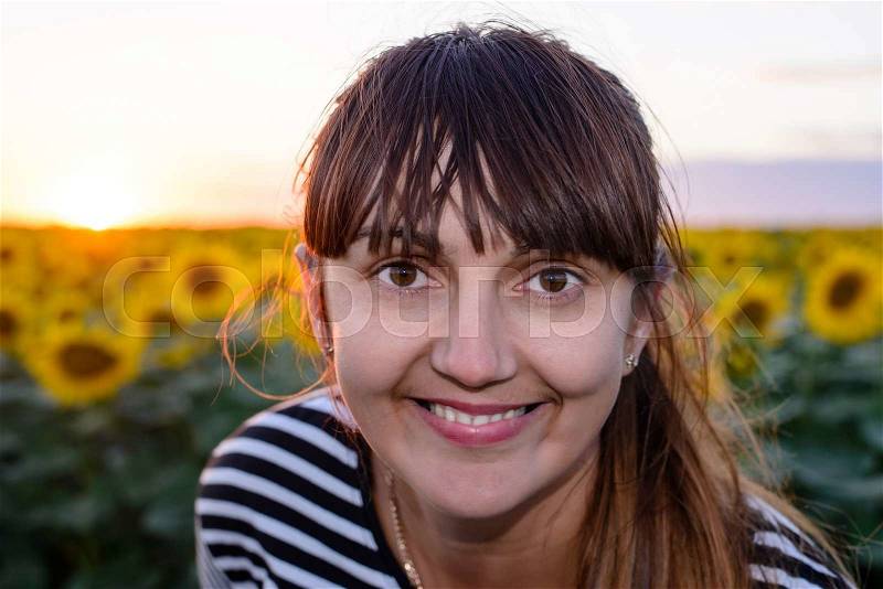 Smiling woman peering into the lens of the camera with a beaming friendly smile against a background of bright yellow sunflowers at sunset, stock photo