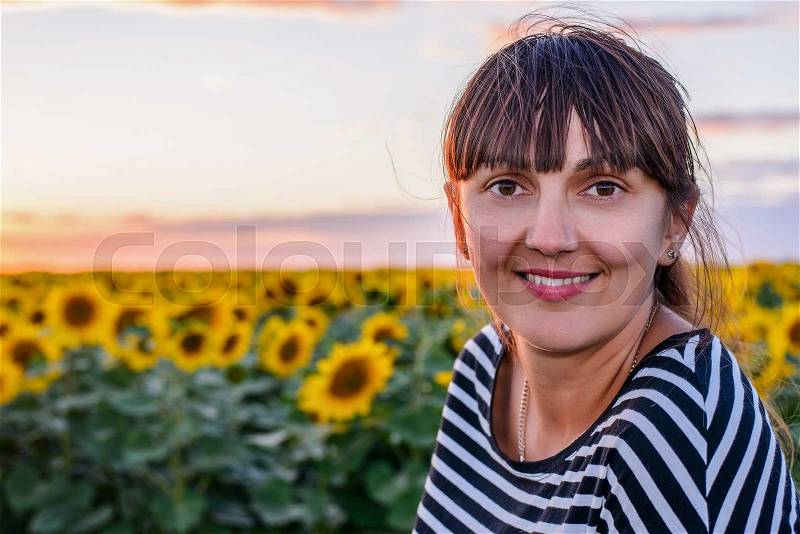 Smiling woman peering into the lens of the camera with a beaming friendly smile against a background of bright yellow sunflowers at sunset, stock photo