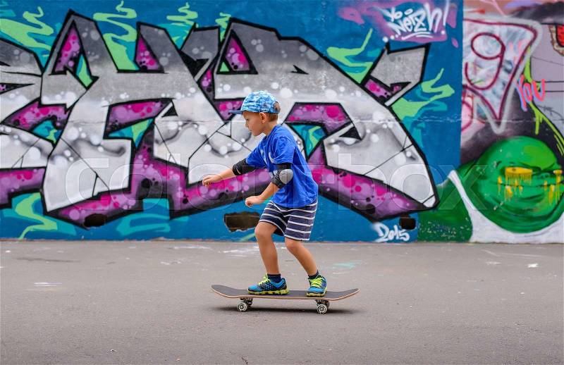 Confident little boy posing on his skateboard in front of a vividly painted graffiti wall with his hands on his hips, stock photo
