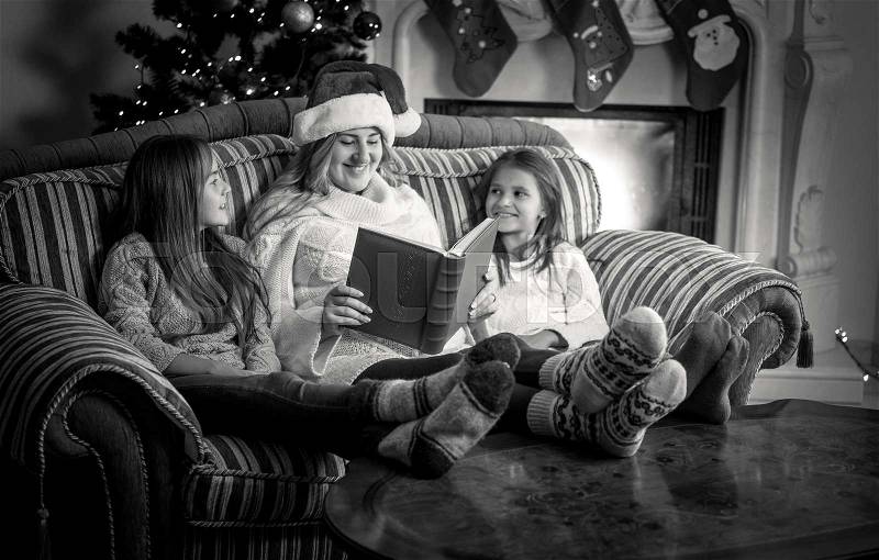 Black and white portrait of mother with two daughter reading book on sofa at fireplace, stock photo