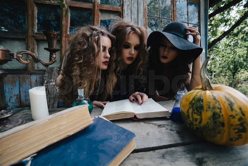 Three vintage witches perform magic ritual, at the table, reading a magical book on the eve of Halloween, stock photo