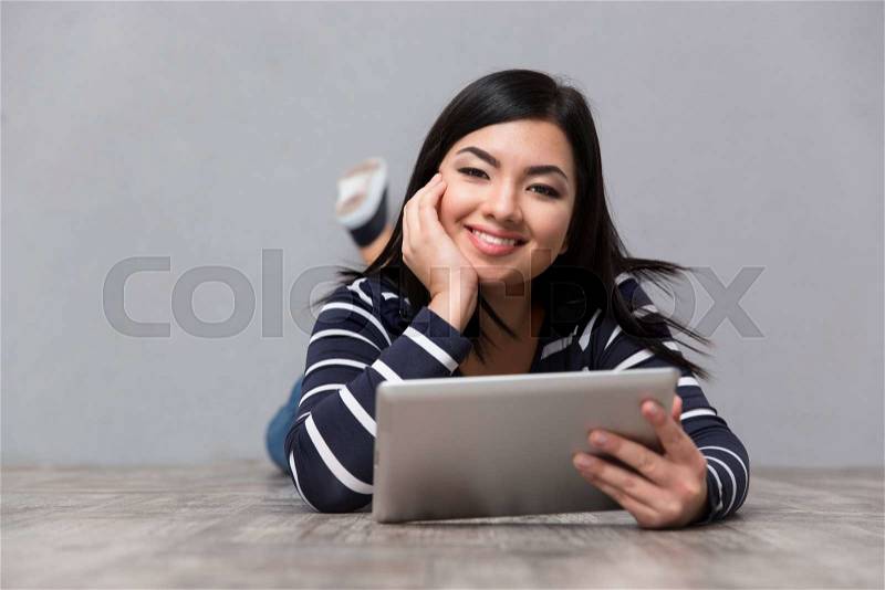 Portrait of a smiling woman lying on the floor with tablet computer on gray background and looking at camera, stock photo