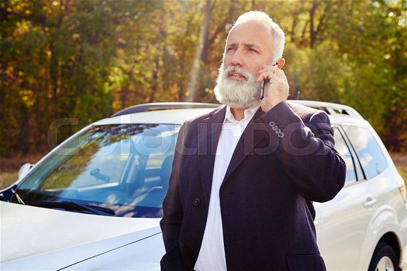 Bearded senior man talking on the phone over white car at outdoor, stock photo