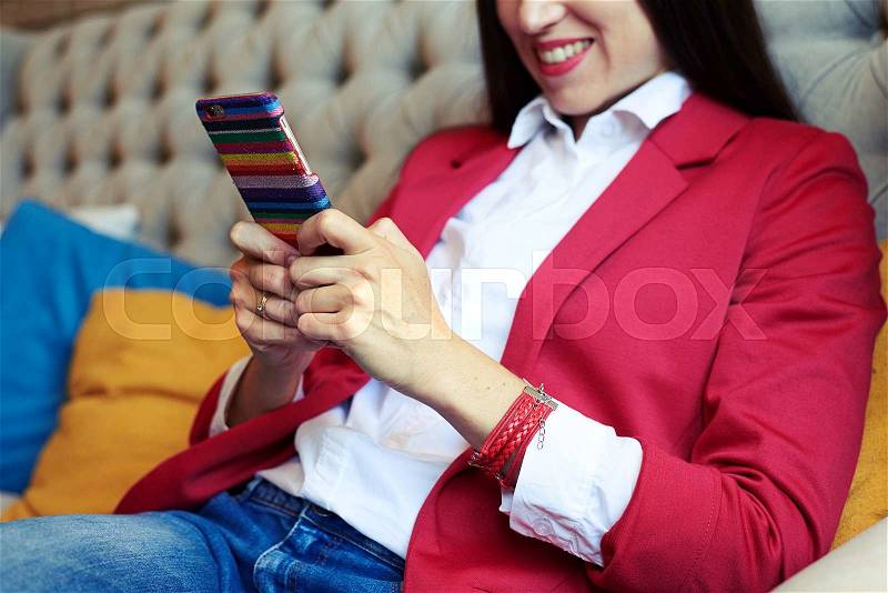 Smiley young woman sitting on sofa and chatting on smartphone, stock photo