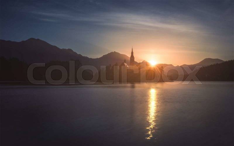 Silhouette of Little Island with Catholic Church in Bled Lake, Slovenia at Sunrise with Castle and Mountains in Background, stock photo
