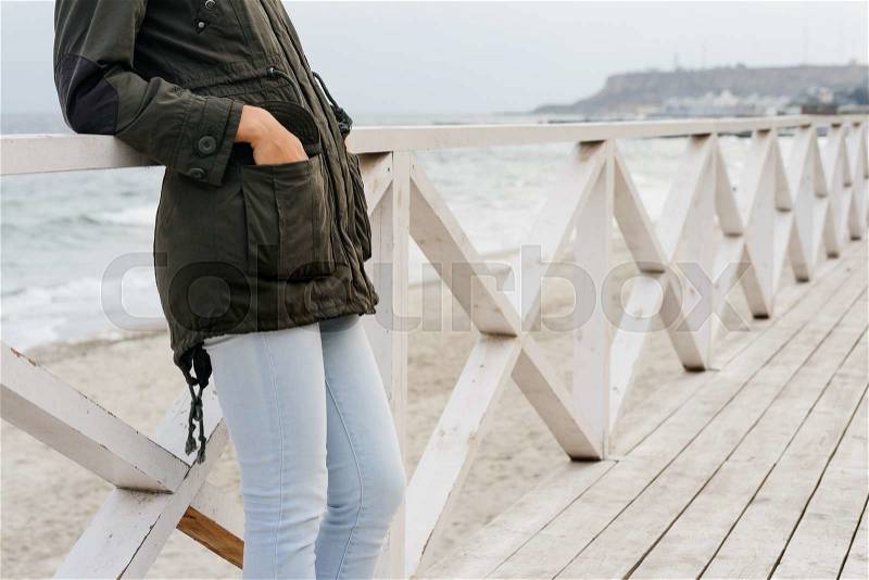 Woman in a green jacket and blue jeans standing on the wooden promenade near the sea. Autumn walk on the beach, stock photo