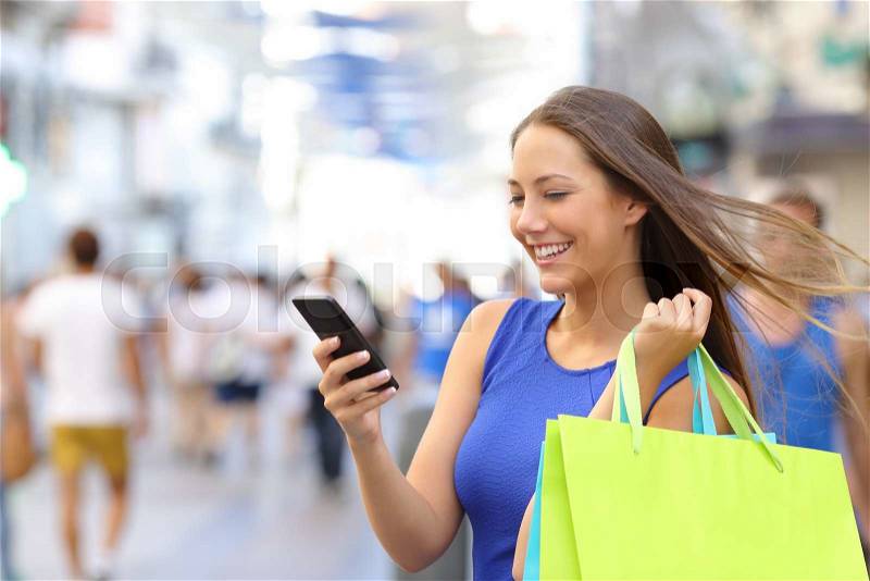 Shopper woman shopping with a smartphone in a commercial street, stock photo