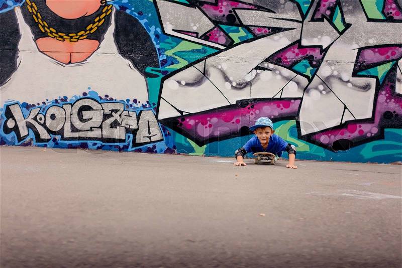 Cute young boy playing on his skateboard using his hands to propel himself towards the camera with a cheeky grin in front of a colorful graffiti wall, stock photo